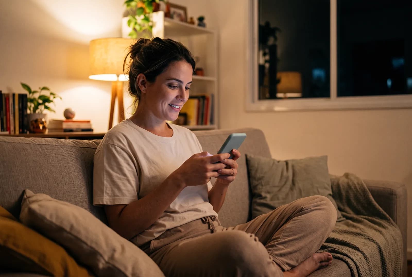Mulher no sofá de casa, à noite, digitando no celular com um leve sorriso.